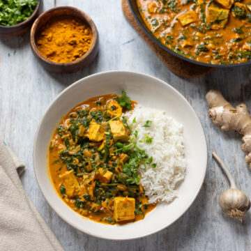 overhead view of a flatly of saag tofu in a bowl with rice and cilantro, with a pan of saag tofu in the corner and small bowls of spices, plus ginger and garlic