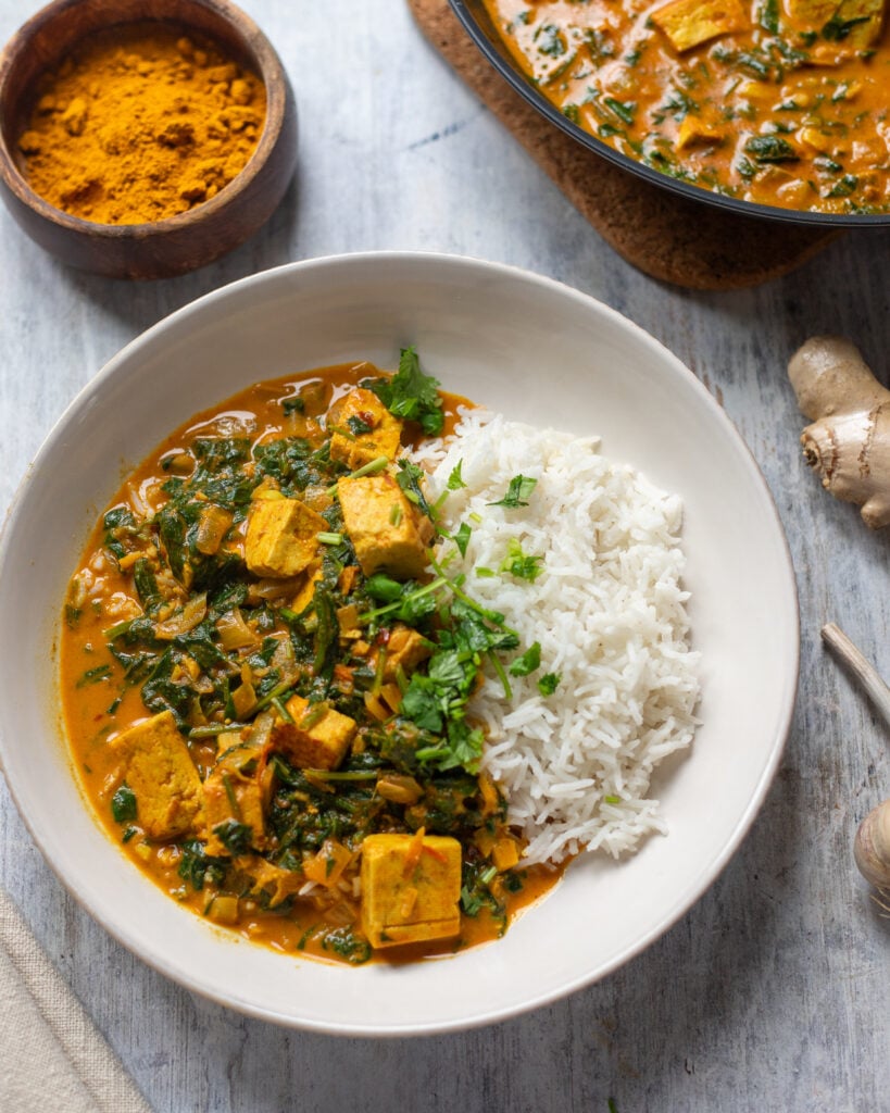 close up overhead view of a flatly of saag tofu in a bowl with rice and cilantro, with a pan of saag tofu in the corner and small bowls of spices, plus ginger and garlic