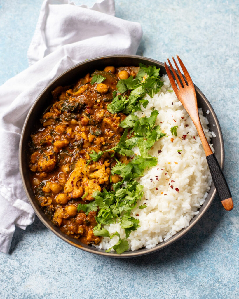 overhead view of a bowl with spicy vegan cauliflower curry with chickpeas, coconut milk, spinach, zucchini and more served with steamed rice and cilantro.