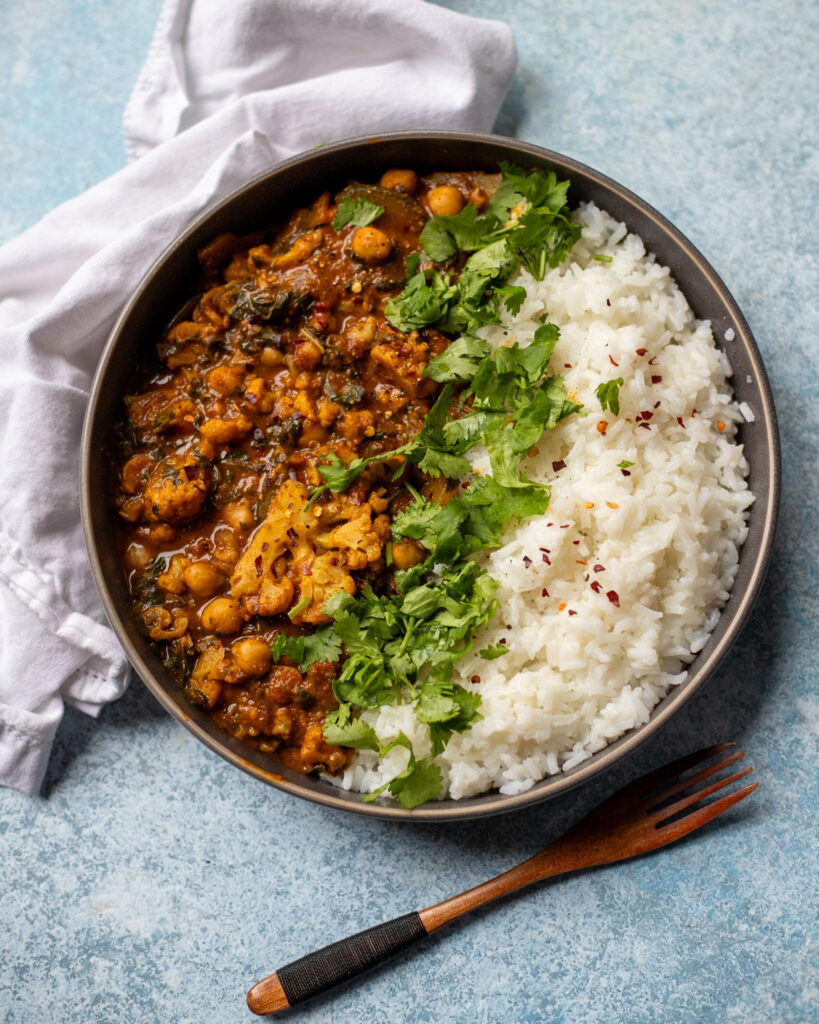overhead view of a gray serving bowl with homemade vegan cauliflower curry with chickpeas in a spicy curry gravy served with white rice, red pepper flakes, and fresh cilantro.