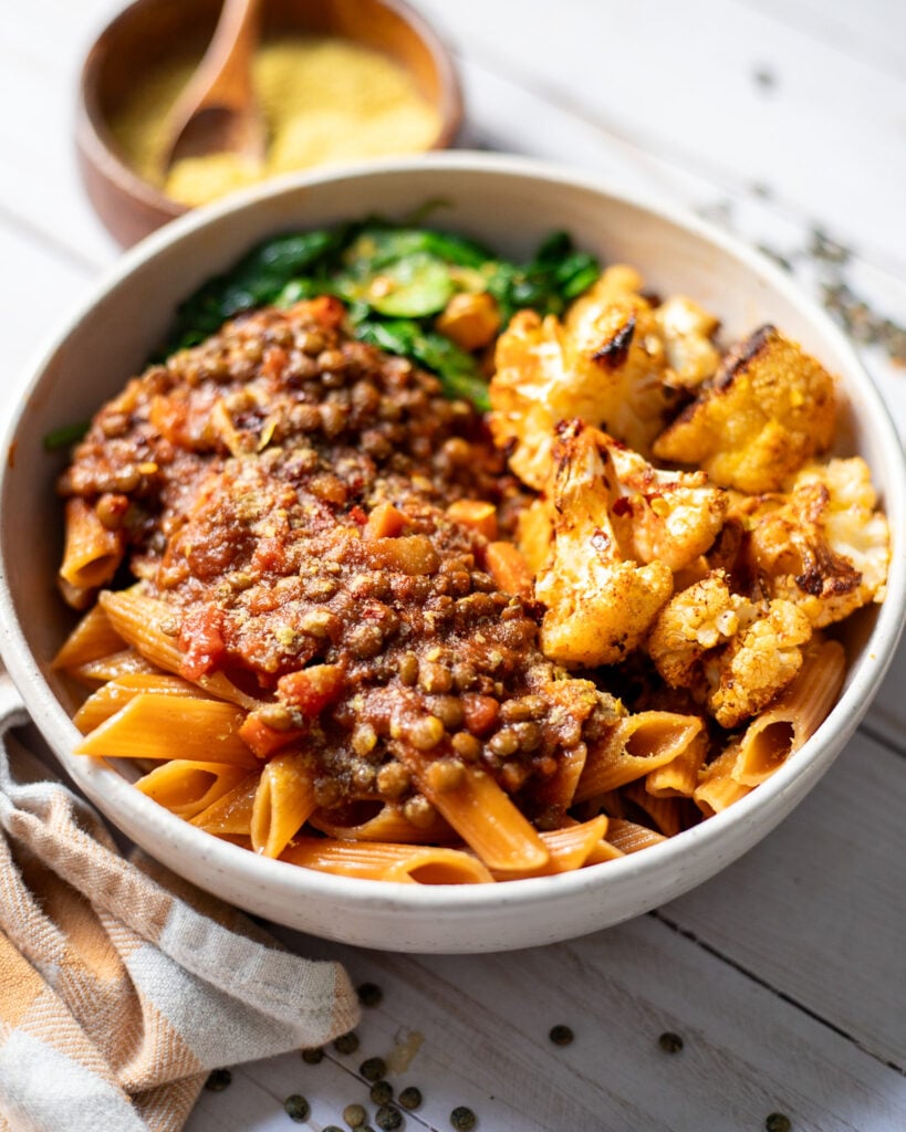 side view of a bowl with vegan bolognese made with green French lentils served with chickpea pasta, roasted cauliflower, and wilted spinach.