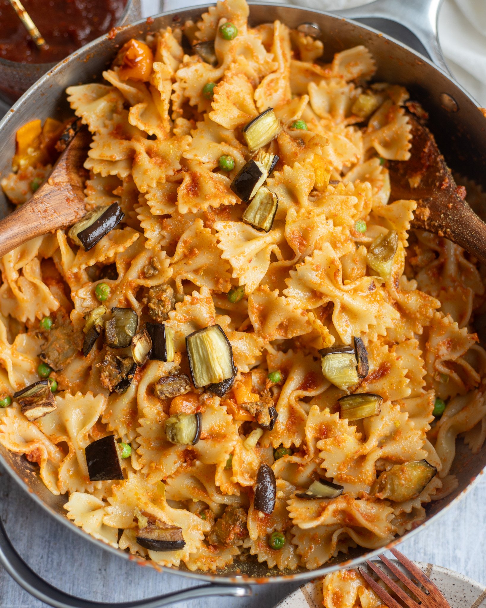 overhead view of a large skillet with farfalle pasta coated in a homemade vegan red pesto pasta with pieces of roasted eggplant, peppers, and peas.