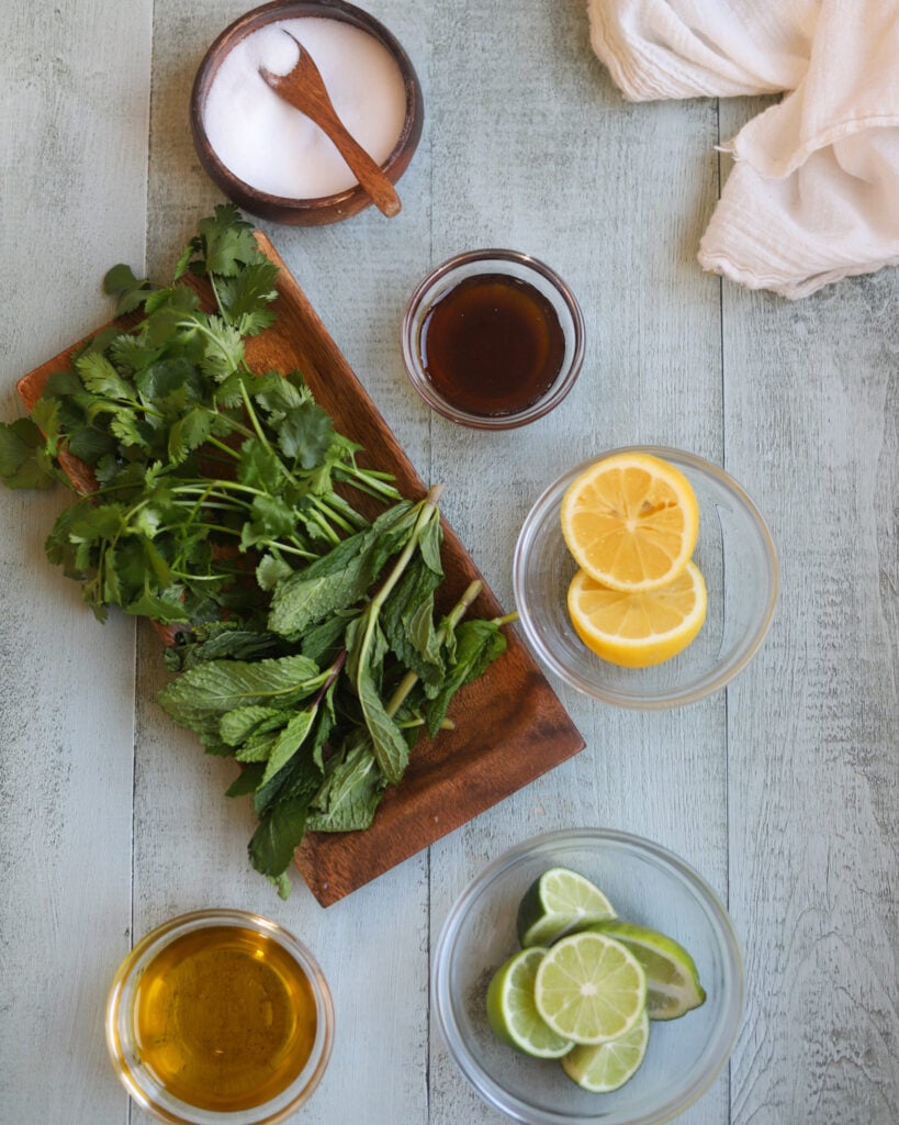 Overhead view of citrus mint salad dressing ingredients including a rectangular wood tray with a large bunch of fresh mint, and round bowls of olive oil, salt, limes, and maple syrup, and two lemon slices