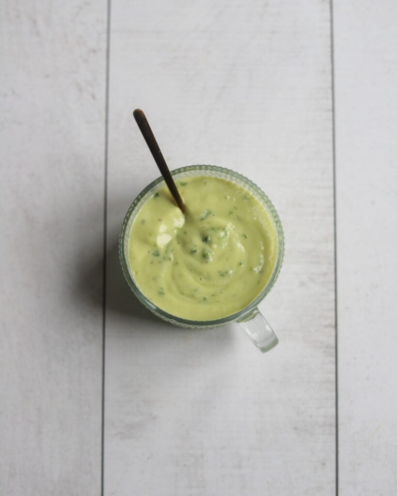 An overhead view of a small round glass bowl of light green creamy avocado lime dressing and a spoon sits alone on a gray surface