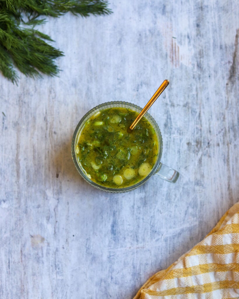 overhead view of a glass serving dish with a spoon filled with green dill Dijon vinaigrette salad dressing sitting on a gray surface along with some fresh dill fronds and a yellow and white checkered cloth.