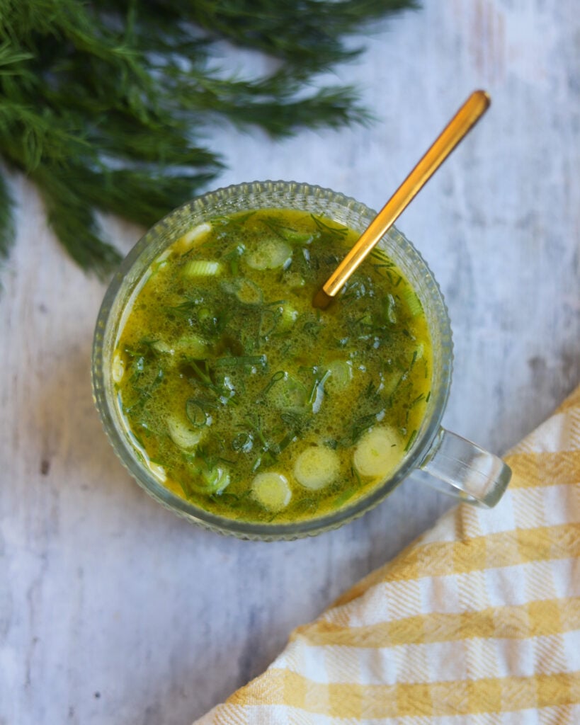 Overhead view of a round glass bowl of dijon and dill salad dressing made with vinegar, olive oil, dill, maple syrup and green onions sits on a gray surface with some fresh dill and a yellow and white cloth