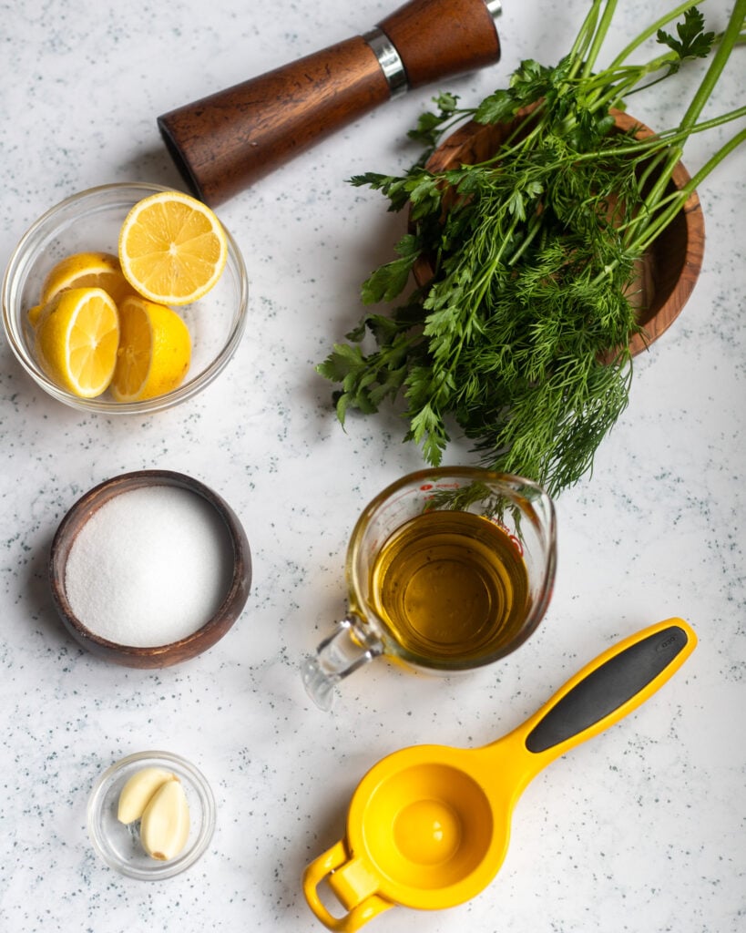 Overhead view of ingredients for garlic herb salad dressing are in small bowls on a gray marble surface, including fresh dill and parsley, olive oil, fresh lemons, salt, garlic and cloves, with a brown pepper mill and a yellow lemon juicer also pictured.