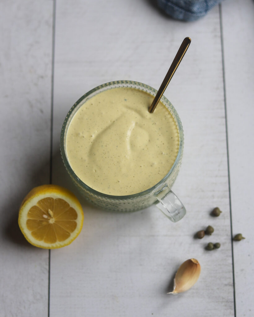 overhead view of a round glass serving bowl of caesar salad dressing made with hempseeds and tofu with lemon juice and olive oil sits on a white surface with a half fresh lemon to the side and a sprinkle of capers and a garlic clove.