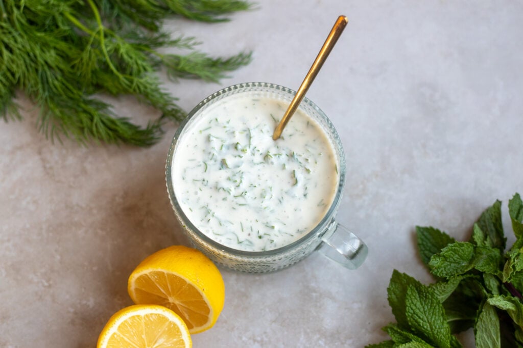 top view of a glass dish of vegan herb salad dressing made with vegan yogurt, dill and mint, lemon juice and maple syrup sits on a gray surface surrounded by fresh dill and mint leaves and a cut open lemon.