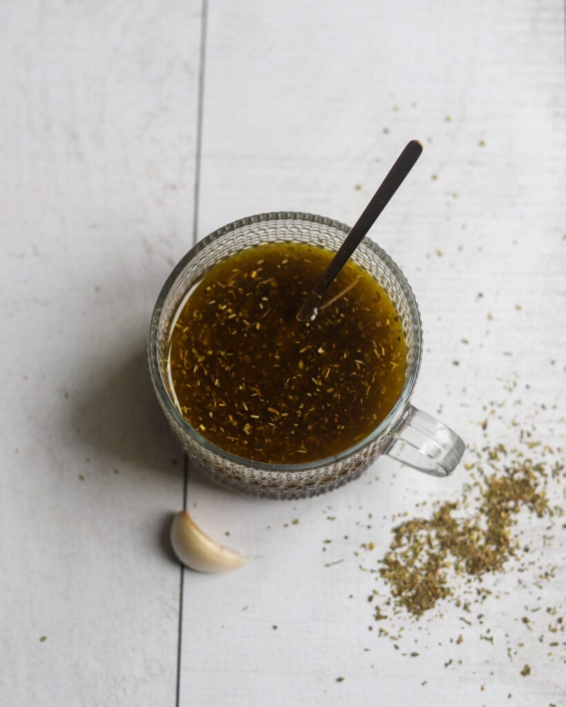 a glass bowl of vegan restaurant-style vegan italian salad dressing is sitting on a white surface with a garlic clove and some italian seasonings scattered nearby.