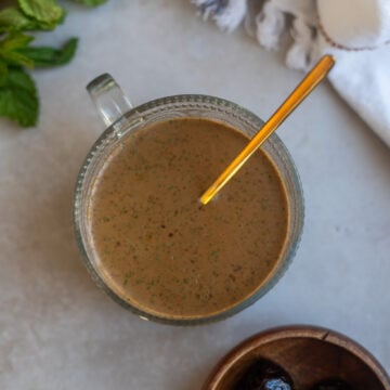 overhead view of a round glass bowl of min and prune dressing with balsamic vinegar, prunes, fresh mint leaves, olive oil and maple syrup on a white surface with a bowl of dried prunes and some sprigs of fresh mint
