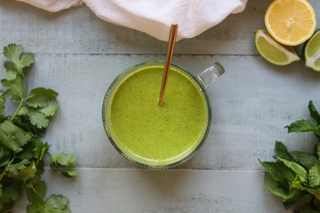 Overhead view of a round glass bowl of light green lemon lime salad dressing with fresh mint, made with olive oil and maple syrup. The bowl is on a gray table along with two bunches of fresh mint and cut lemons and limes.