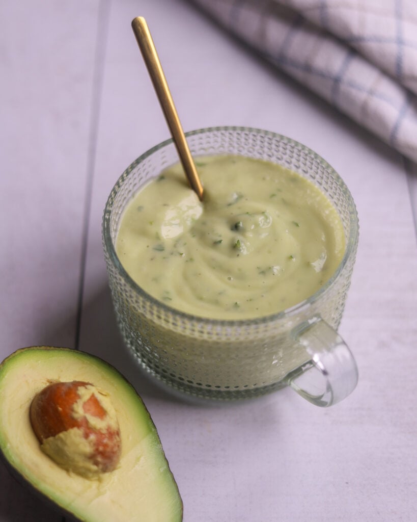 side angle view of a glass serving dish of tangy avocado ranch dressing sits on a gray surface with a half avocado and a checkered dish towel