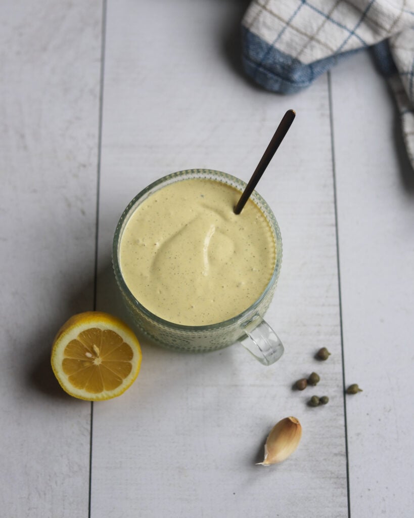A bowl of vegan tofu-based caesar dressing is sitting on a white surface along with half of a fresh lemon, s few capers and a garlic clove