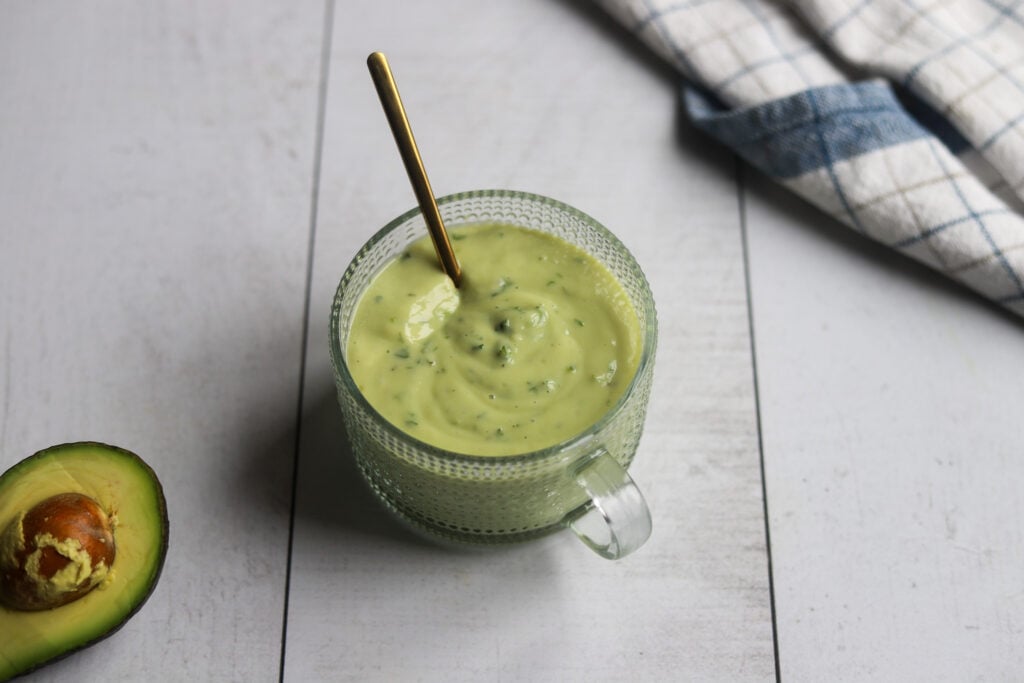 a round bowl of vegan avocado lime dressing sits on a gray surface with half an avocado and a blue and white checkered dish cloth in the corner of the image