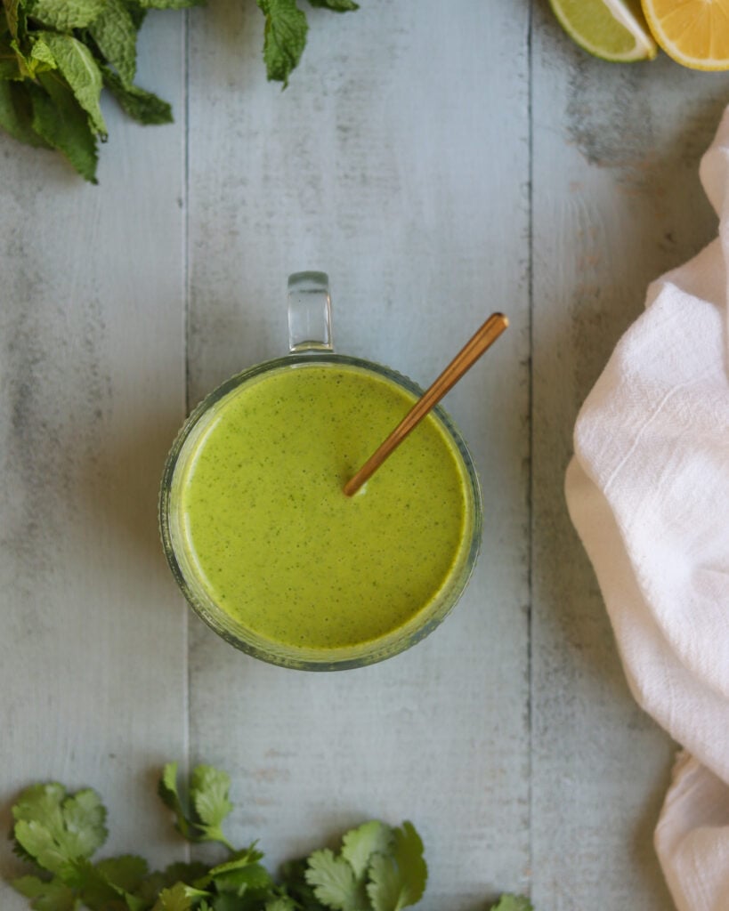 Overhead view of a small bowl of vegan citrus salad dressing on a table surrounded by bunches of fresh mint. Ingredients include sweet maple syrup, lemon and lime juice and fresh mint leaves in an olive oil base.