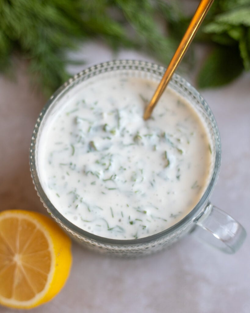 closeup of a top view of a bowl of vegan herby yogurt dressing made with lemon juice, herbs, yogurt and spices is on a gray surface with a half of a fresh lemon and some dill and mint in the background.