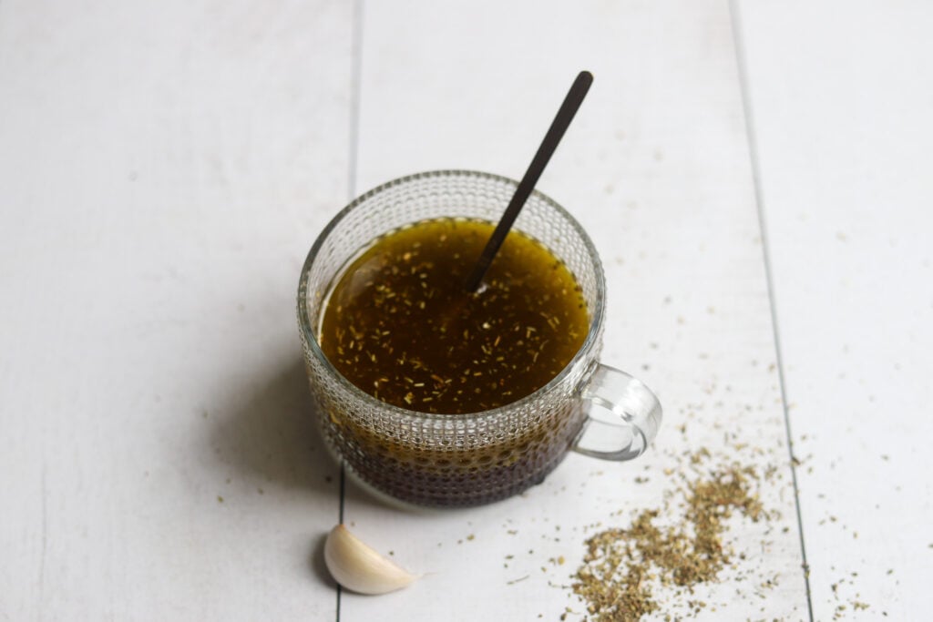 side view of a bowl of red wine vinegar Italian salad dressing on a white surface with fresh garlic and italian herb seasoning.
