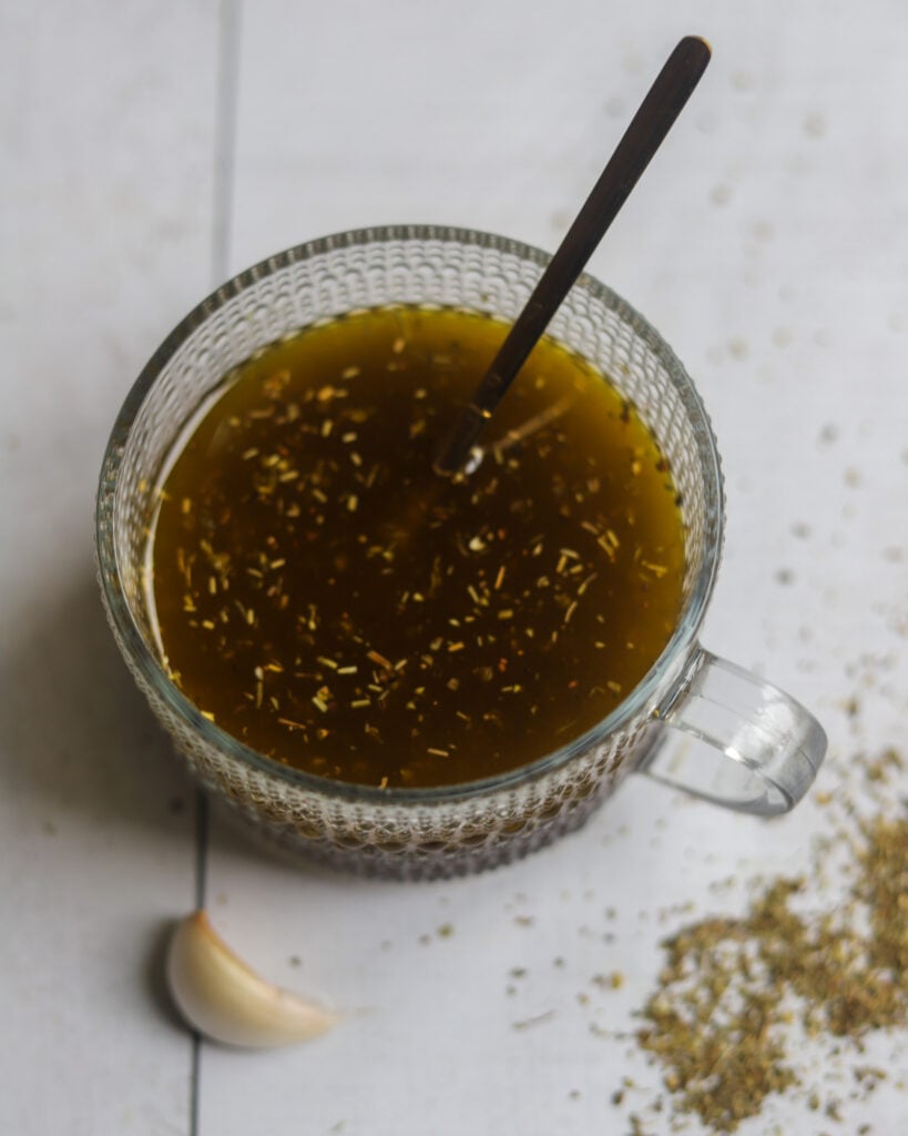 overhead view of a glass serving dish with brown Italian style vegan salad dressing made with red wine vinegar and maple syrup, next to a fresh garlic clove and scattered italian dry seasonings.