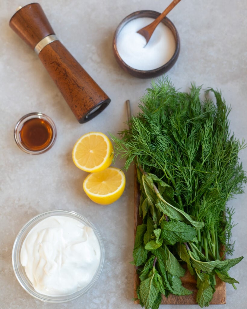 overhead view of a gray surface on which sit the ingredients to make creamy yogurt herb salad dressing including vegan yogurt, fresh lemons, maple syrup, fresh dill and mint, salt and pepper.