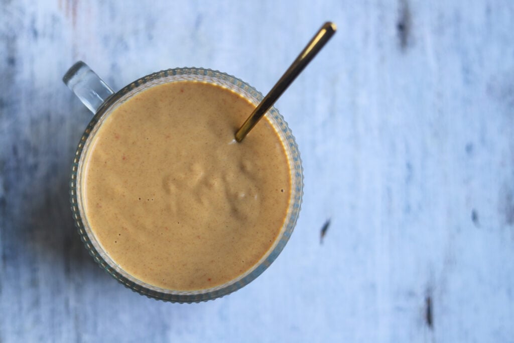 closeup overhead view of a glass bowl with serving spoon, the bowl filled with smooth light brown healthy lime peanut butter dressing that is made from soy sauce, sweet chili sauce, rice vinegar, juicy fresh limes and peanut butter sitting on a gray marbled surface.