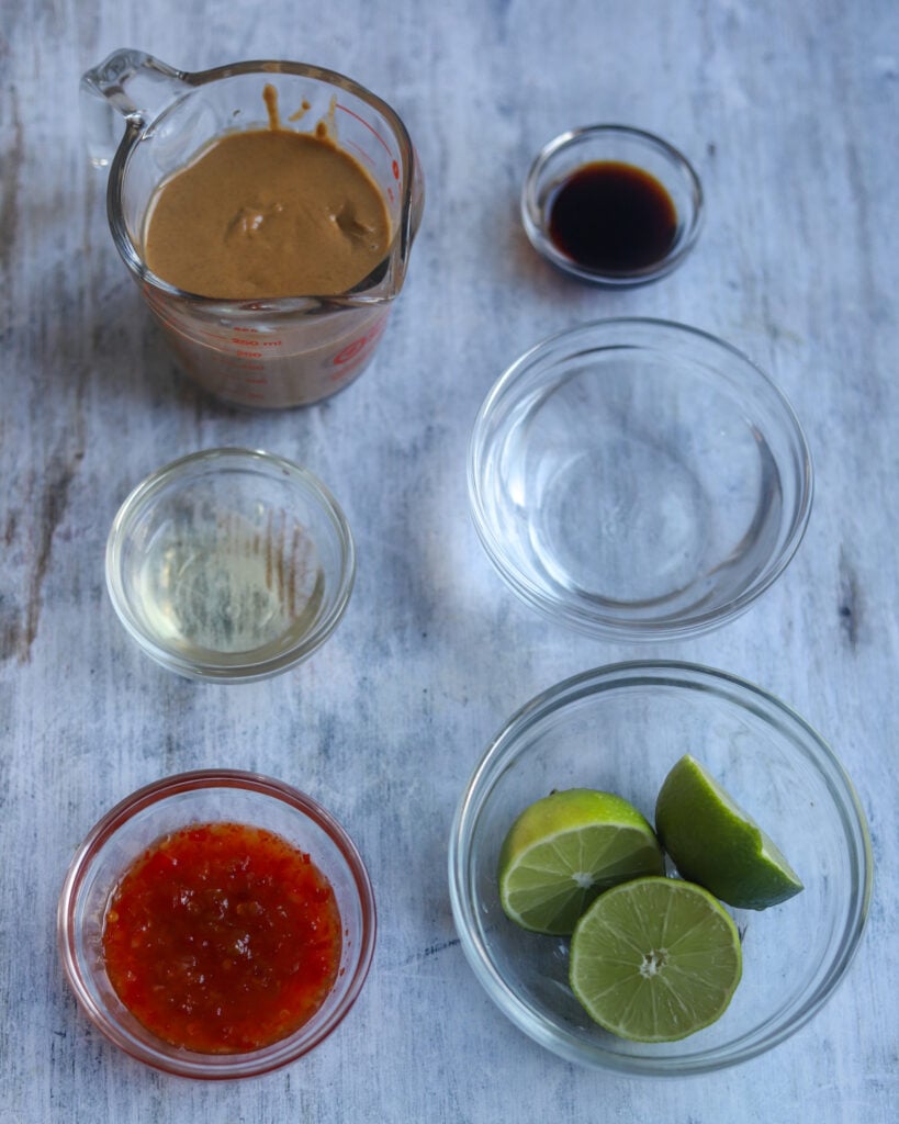 overhead view of several glass bowls of various sizes holding ingredients for peanut dressing like fresh cut limes, rice vinegar, red sweet chili sauce, water, soy sauce and a glass measuring cup full of smooth peanut butter all sit on a gray surface.