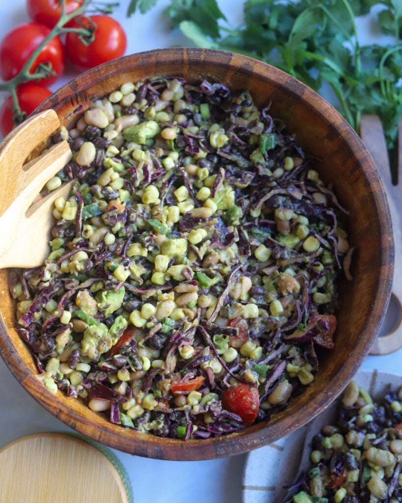 An overhead view of a wooden bowl filled with southwest 3 bean salad made with pintos, white and black beans, corn, tomatoes, fresh cilantro, jalapeno and avocado, with a bowl of lime cilantro salad dressing, some tomatoes fresh cilantro leaves and a plate of salad around the bowl.