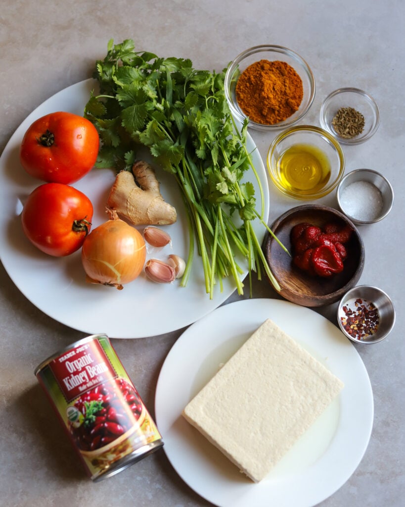 Ingredients for Rajma recipe overhead view: tomatoes, onion, garlic, ginger, kidney beans, tofu, curry powder, tomato paste, salt, cumin seeds, red pepper flakes and extra virgin olive oil