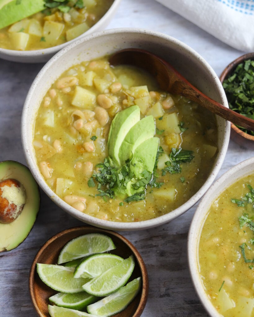 Bowl of Hatch Green Chili topped with avocado and cilantro with limes and chopped cilantro on the side