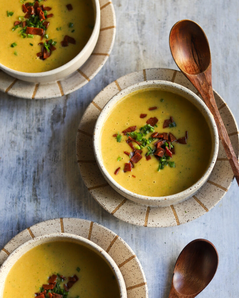 overhead view of 3 bowls of broccoli cheddar soup with wooden spoons on the side and bacon and chive garnish on top