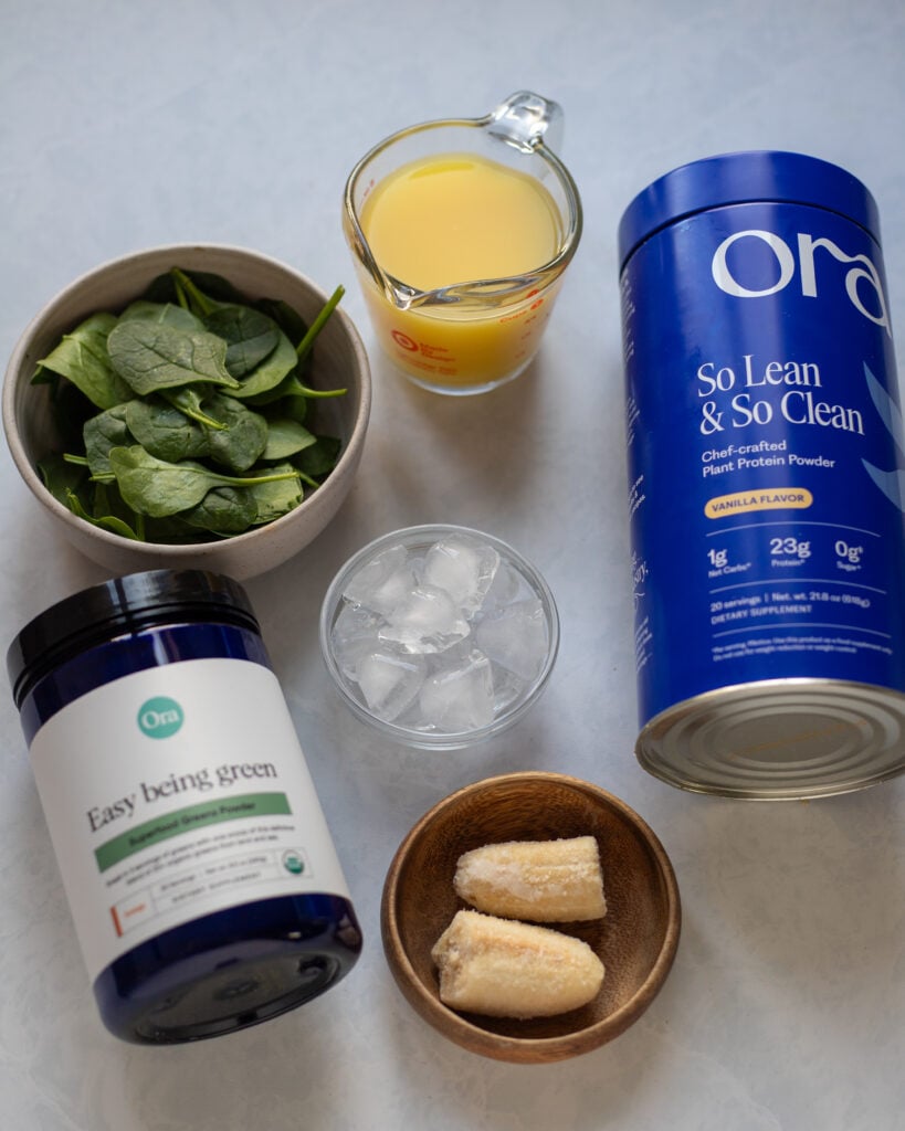 Ingredients for healthy green smoothie with vegan protein powder. frozen banana, greens powder, spinach, orange juice, water, ice and protein powder