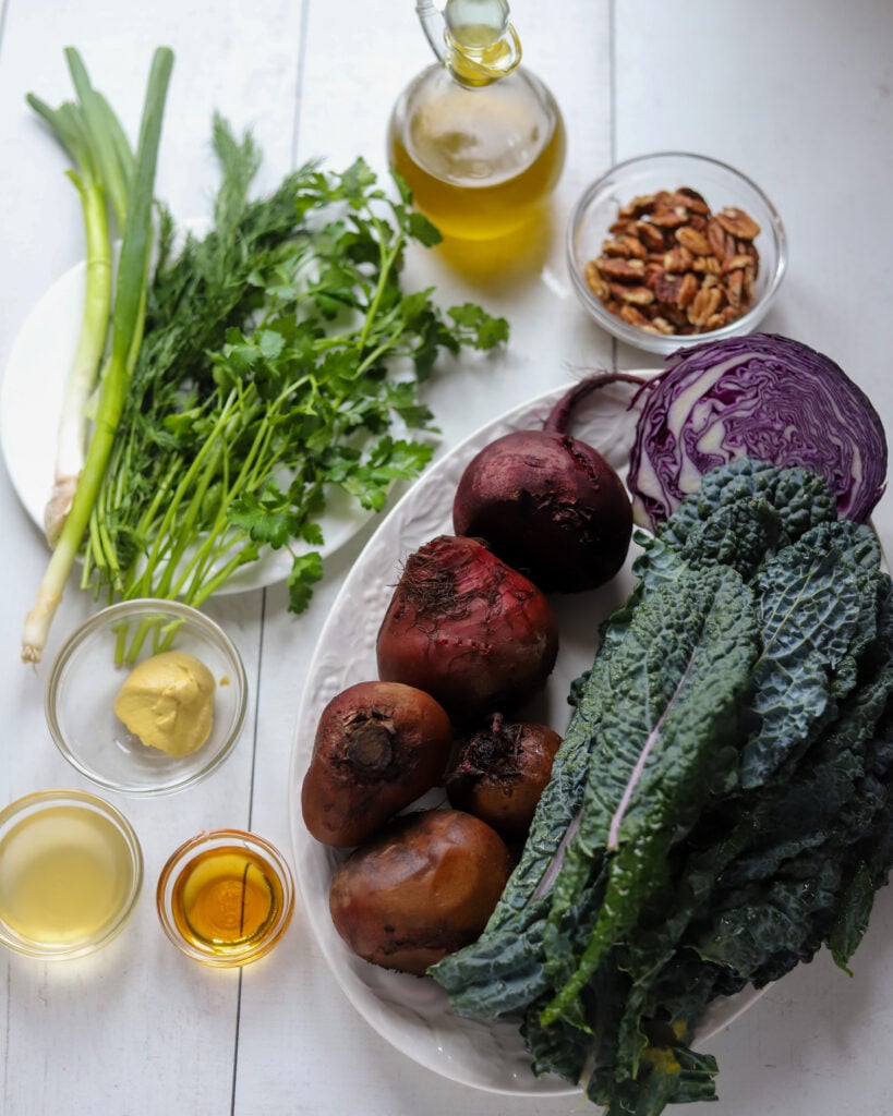 ingredients for beet kale salad laid out in a flatly: kale, beets, green onions, parsley, dill, cabbage, pecans oil, vinegar, dijon mustard and maple syrup
