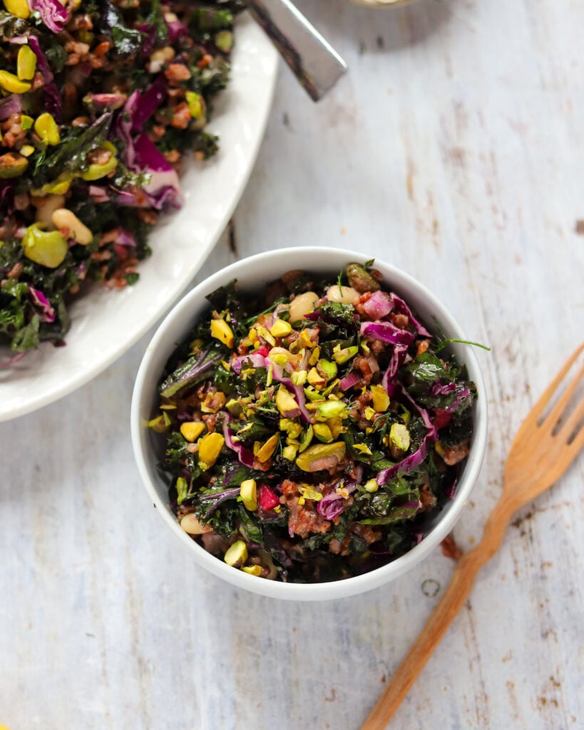 Overhead view of a small bowl of kale and rice salad, there are purple bits of cabbage and radishes, chopped green olives, beans and pistachios visible.