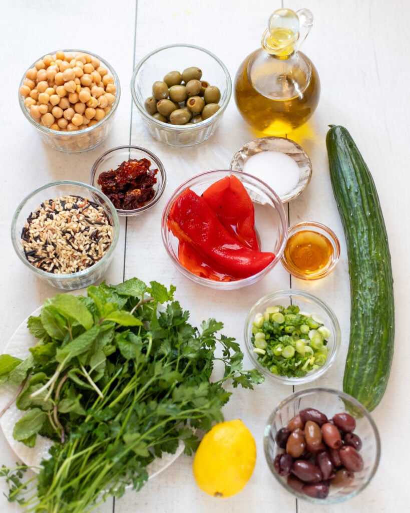 Overhead view of Mediterranean Wild Rice Salad ingredients laid out in small bowls Chickpeas peppers, rice, green onions, herbs, lemon, olive oil, maple syrup and Olives in a wood bowl with serving spoons