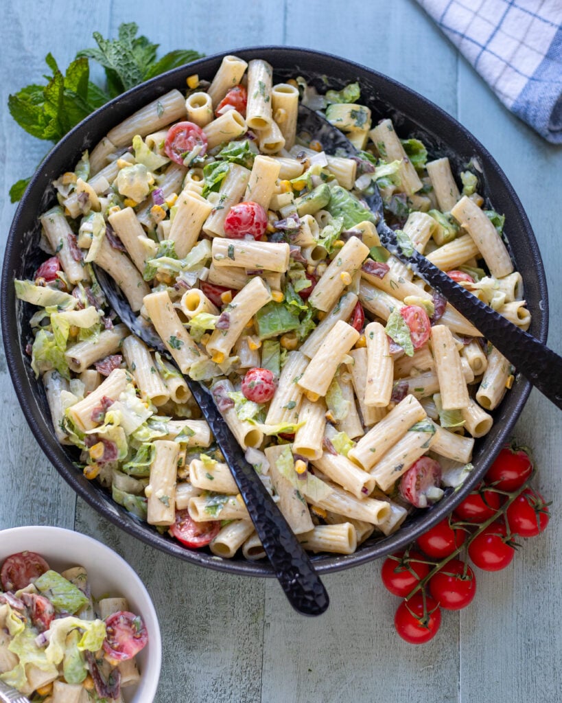 Overhead view of a large bowl of vegan BLT pasta salad with serving utensils, plus a small bowl of pasta salad, herbs and cherry tomatoes