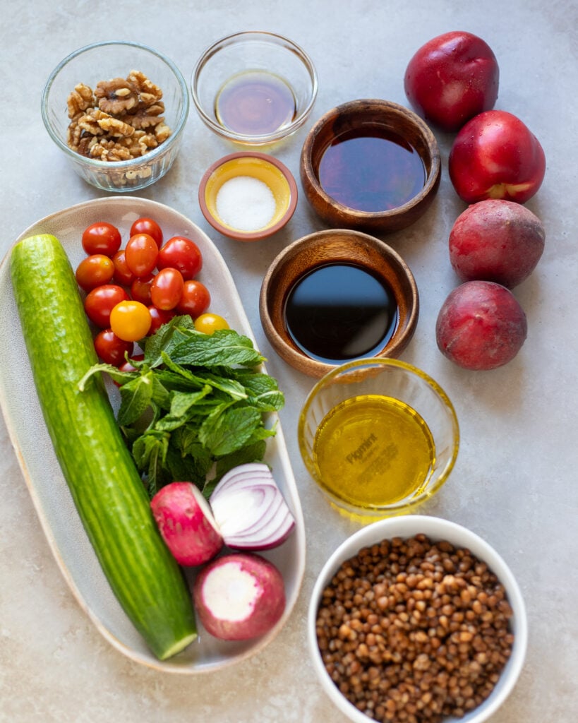 ingredients for summer peach salad with lentils in a flat lay with cucumber, cherry tomato, mint, lentils, walnuts peaches and nectarines, extra virgin olive oil, red wine vinegar, balsamic vinegar, pure maple syrup and extra virgin olive oil